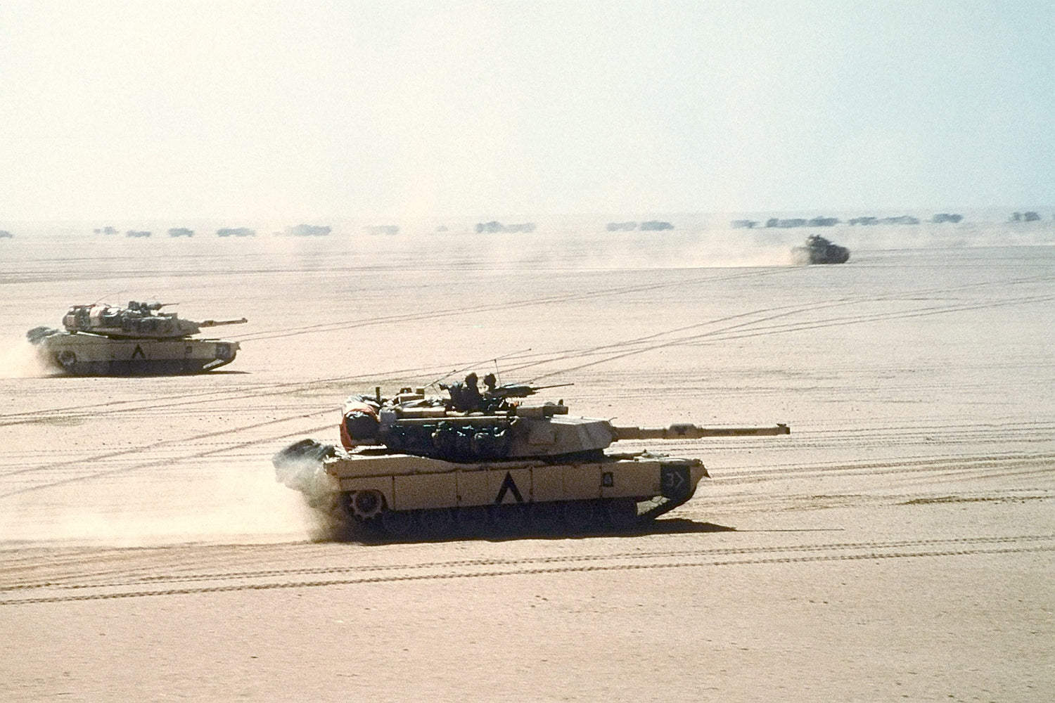 Abrams move out on a mission during the Gulf War. A Bradley IFV and logistics convoy can be seen in the background.
