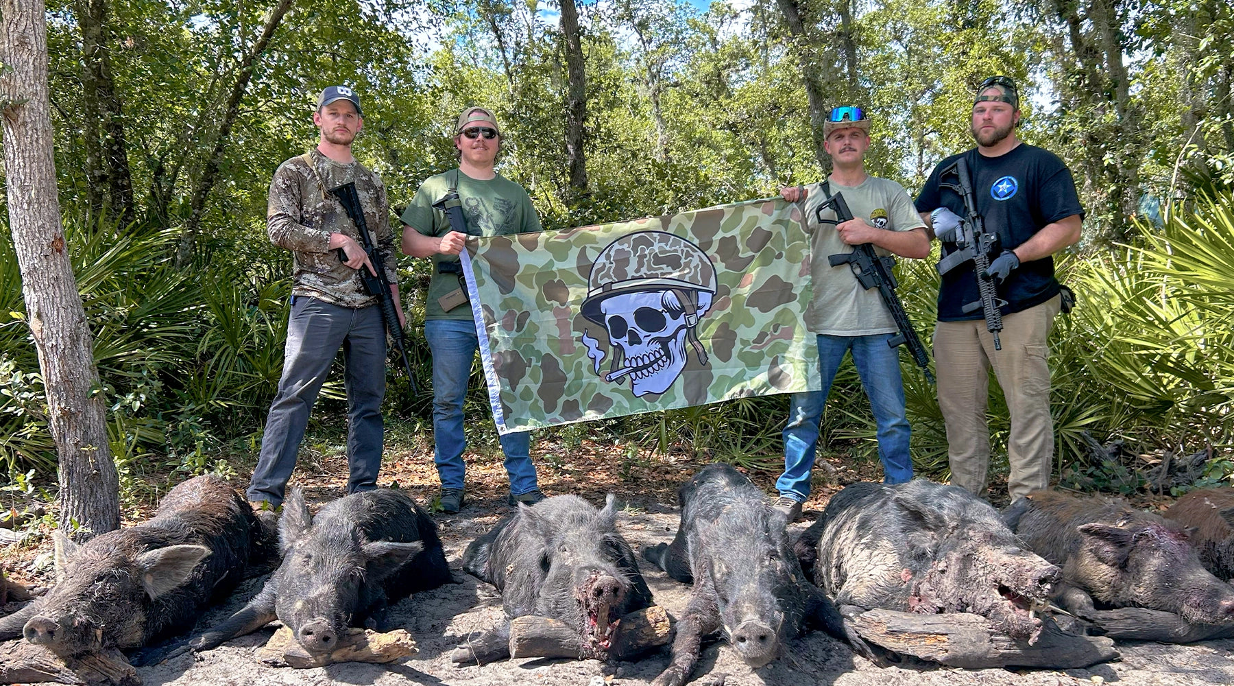 Owners of Tacticallyacquired.com holding a camouflage flag with a skull design, surrounded by hunted wild boars in a forest setting.